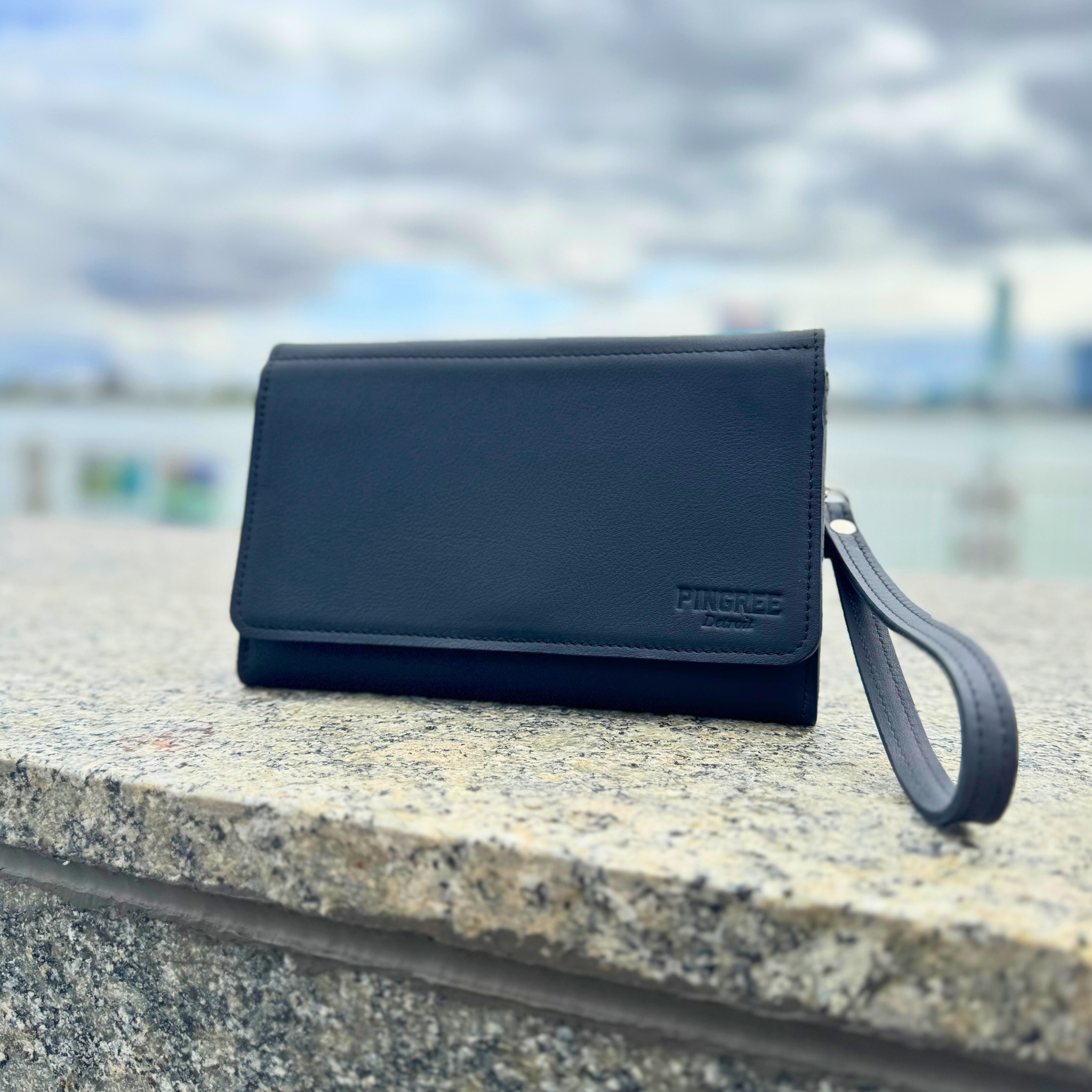 The Adeline Lux, pictured here with the Detroit rver in the background, exterior all black leather, upcycled from the automotive industry, with clean rounded corners, magnetic closure, and a wristlet. This 5-in-1 bag is handmade in Detroit, MI USA, with upcycled leather from the automotive industry. Each comes with a picture of the Veteran or Detroiter who made it.