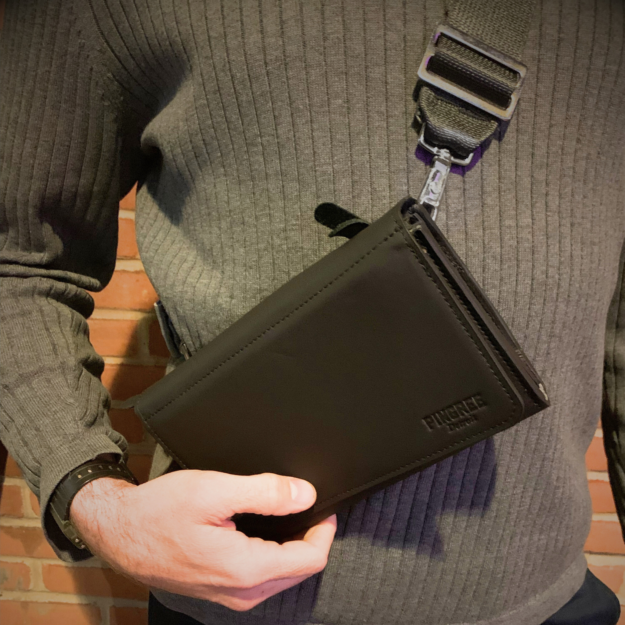 The Adeline Lux, pictured here as a crossbody on a male model, with seatbelt straps, a stone grey interior, and exterior all black leather, upcycled from the automotive industry, with clean rounded corners, magnetic closure, and a wristlet. This 5-in-1 bag is handmade in Detroit, MI USA, with upcycled leather from the automotive industry. Each comes with a picture of the Veteran or Detroiter who made it.