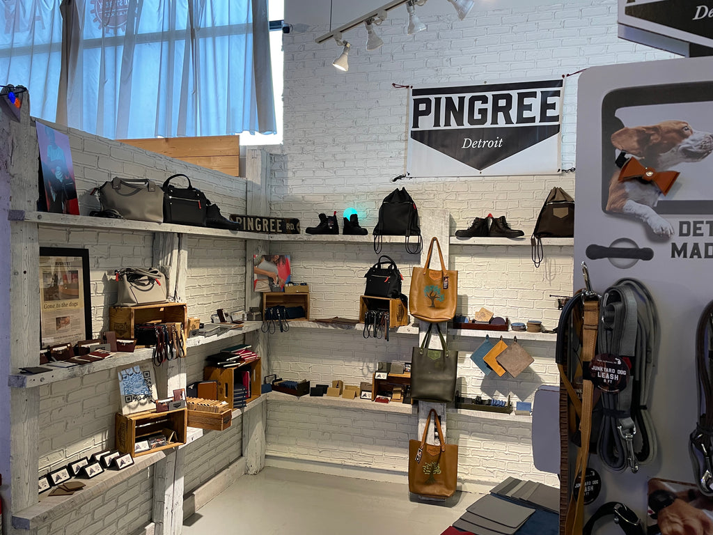 Handmade leather goods displayed on a table including a yellow leather tote, drawstring backpack in black, all made in Detroit by Veterans and Detroiters. There is a Pingree sign and it's at Eastern Market and Rust Belt in Detroit. All handmade sustainable leather 