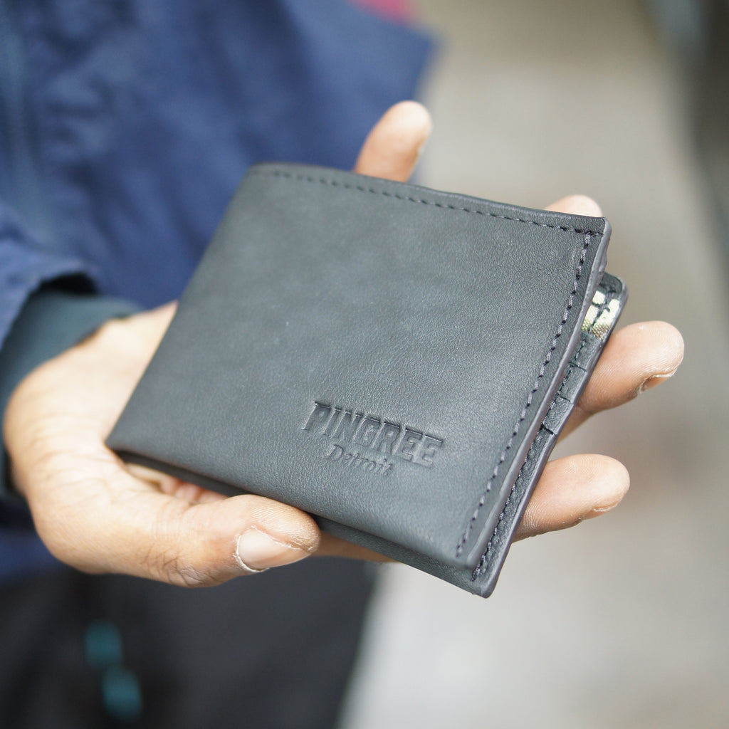 Onyx black billfold wallet, pictured here closed and in a hand. Made in Detroit, Michigan USA. Big enough for George Costanza, 10 pockets, leather upcycled from the production of new cars, handmade in Detroit by U.S. Veterans and Detroiters. Liner made from upcycled material from the production of Army tents.  