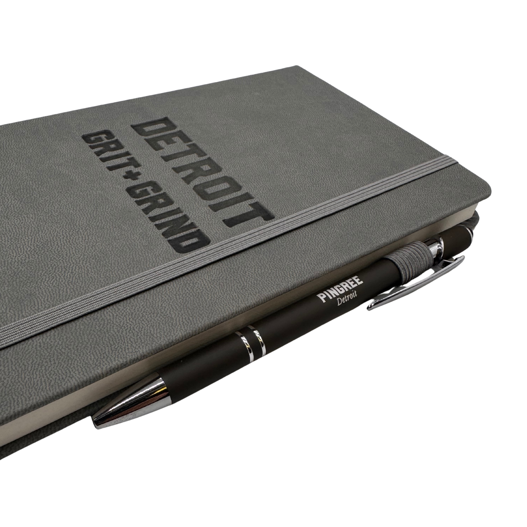 Gray notebook with 'Detroit Gut Grind' text and a pen on a white background. Our hand assembled Journal crafted with 100% recycled paper pulp with lined pages. Detroit made me. Includes a pen, double bookmarks a front pocket and a rear pocket and elastic closure that we sew for reinforcement. A 5 size, comparable to a moleskine style, fits inside of our portfolio or works great as a standalone.