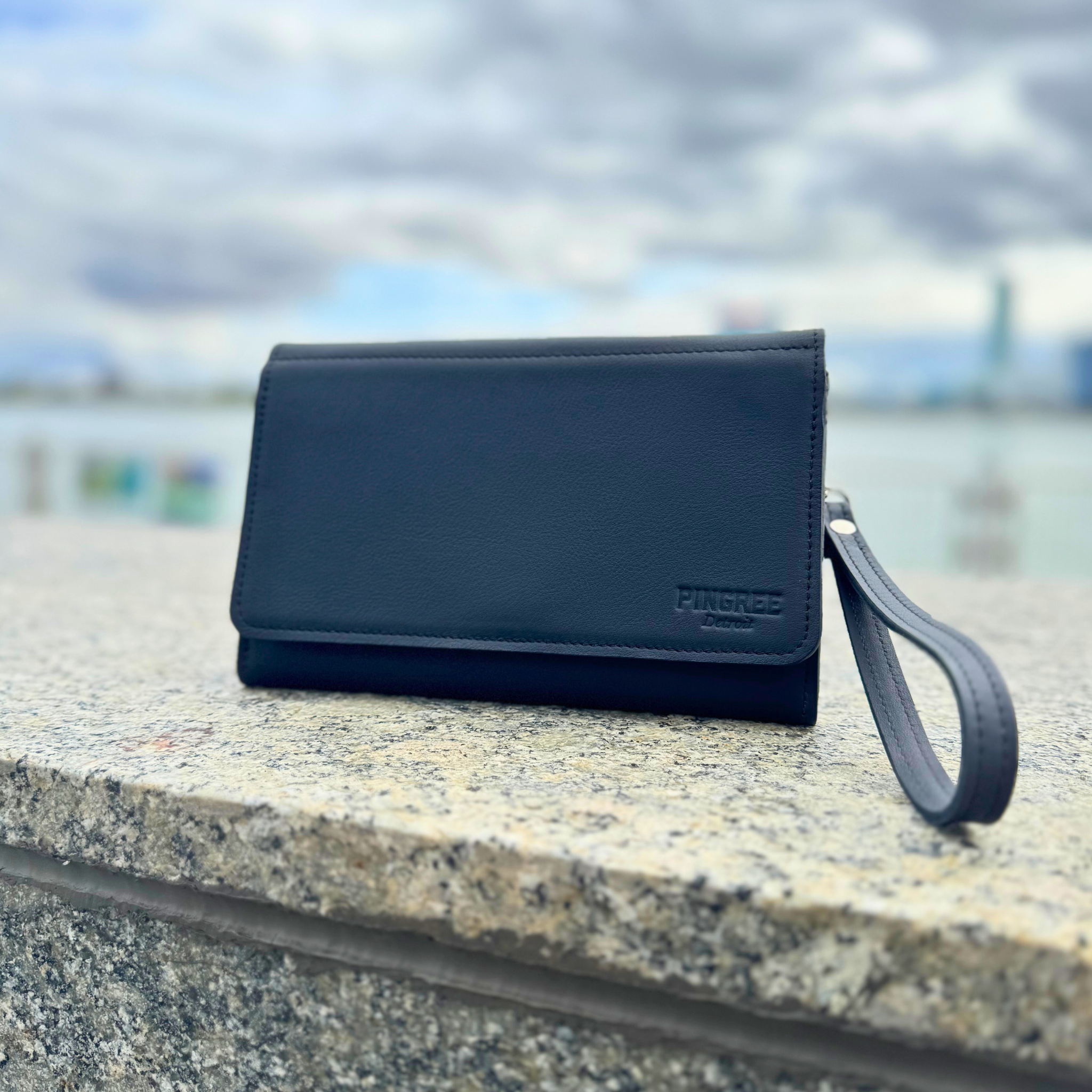 The Adeline Lux, pictured here with the Detroit rver in the background, exterior all black leather, upcycled from the automotive industry, with clean rounded corners, magnetic closure, and a wristlet. This 5-in-1 bag is handmade in Detroit, MI USA, with upcycled leather from the automotive industry. Each comes with a picture of the Veteran or Detroiter who made it. 