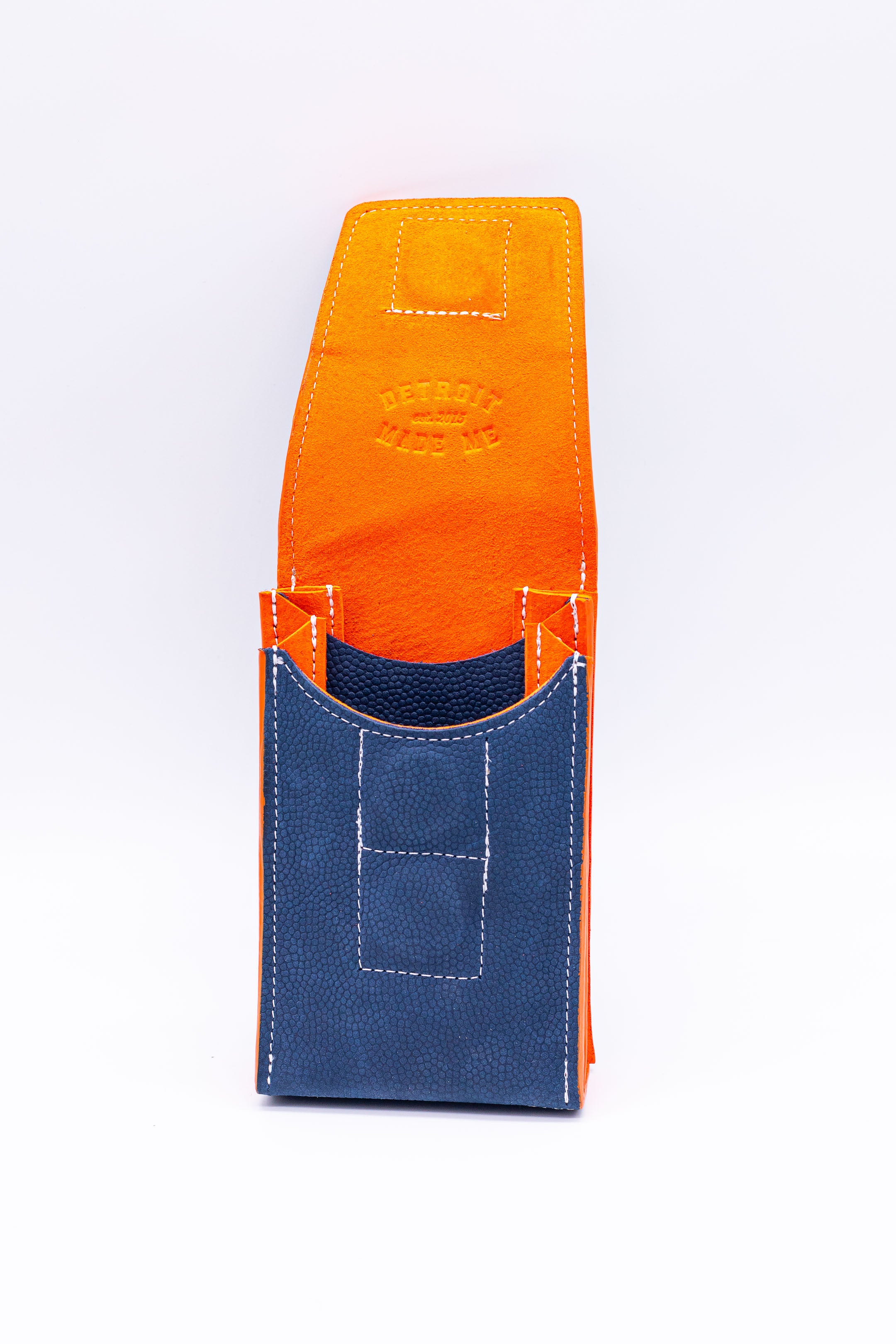 Navy blue pebbled leather on the exterior with orange on the inside and edges. A stadium bag that can be worn on a belt or as a crossbody. Carries your phone and features magnetic closure.  With leather strap on a white background. Made in Detroit with leather upcycled from the automotive industry with the Pingree logo on the front and silver hardware made in America. 