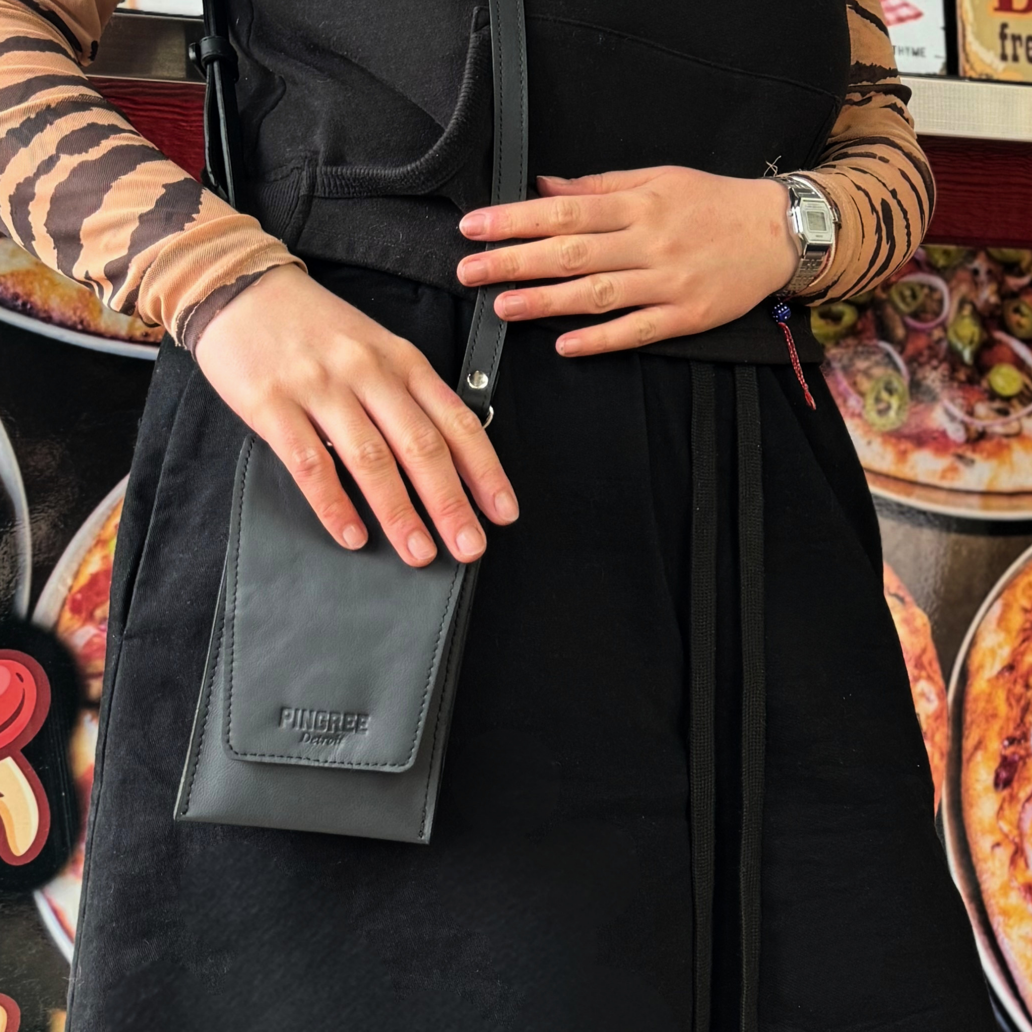 A woman wearing a black dress and tiger-print sleeves carries a black leather crossbody phone bag with a flap pocket. The brand name 'Pingree Detroit' is embossed on the front of the bag. the MVB.