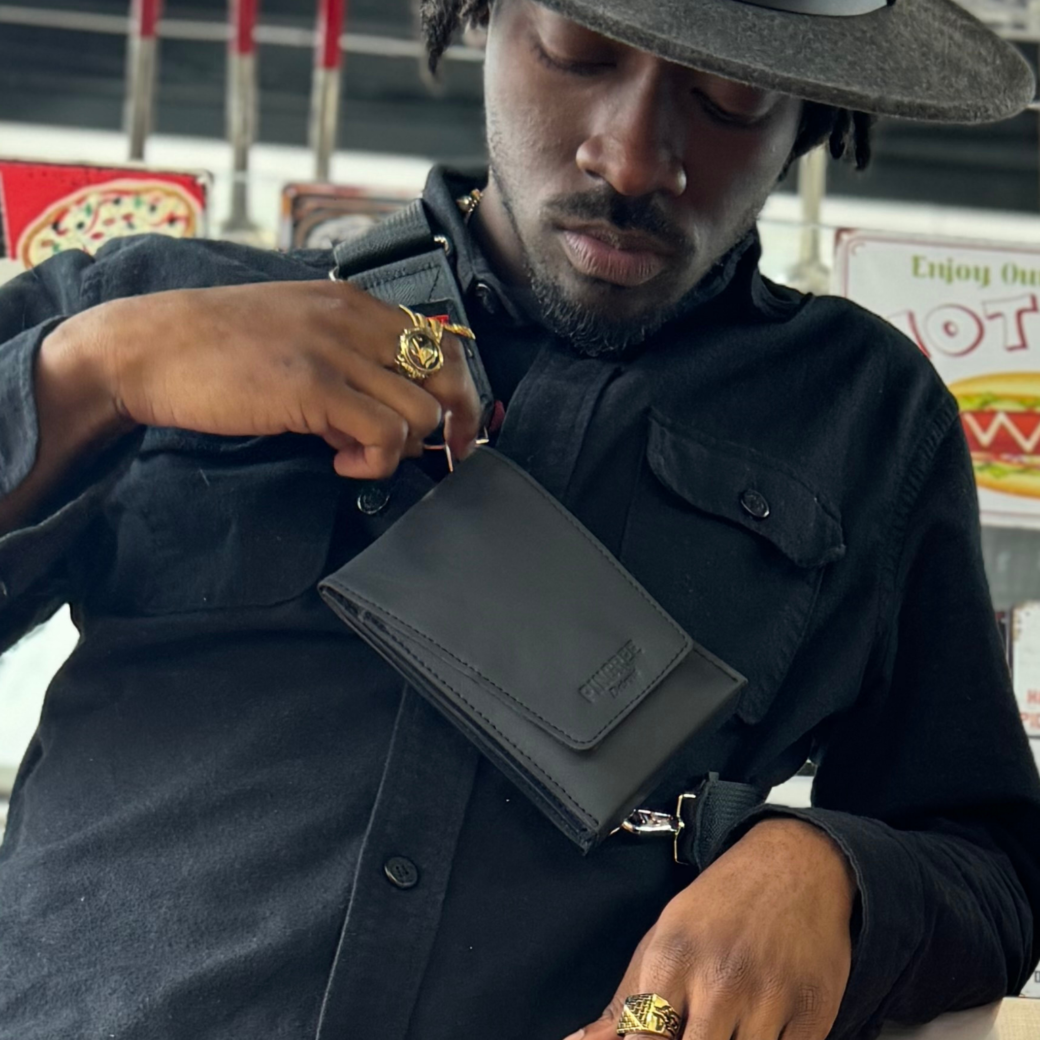 A man wearing a black shirt and hat demonstrates the use of a black leather crossbody wallet with the wallet (Motown Venue Bag) hanging at his waist.